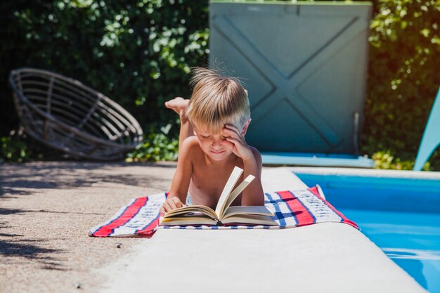 Young boy reading book lying poolside