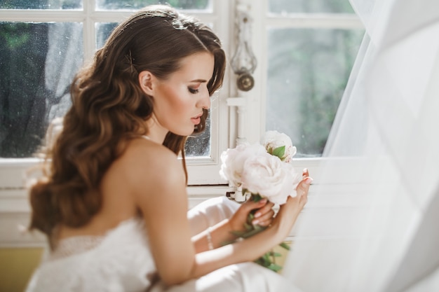 Free photo young bride holds a bouuqet of peonies siting on the windowsill in the bright morning