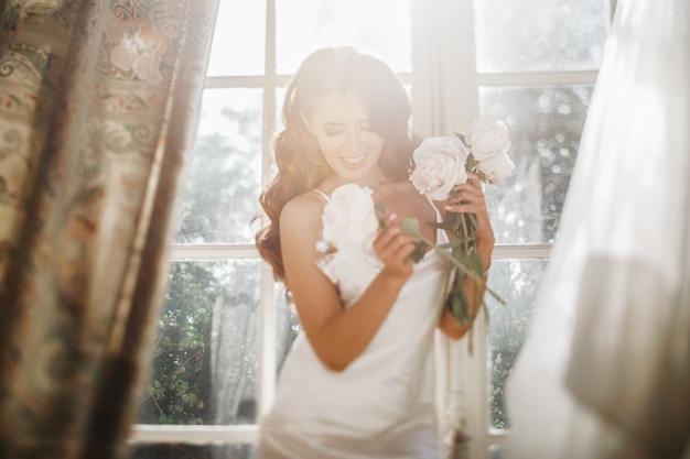 Free photo young bride holds a bouuqet of peonies siting on the windowsill in the bright morning