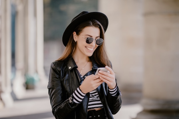 Young brown-haired girl in a leather jacket, black hat on the city promenade and plays on mobile phone