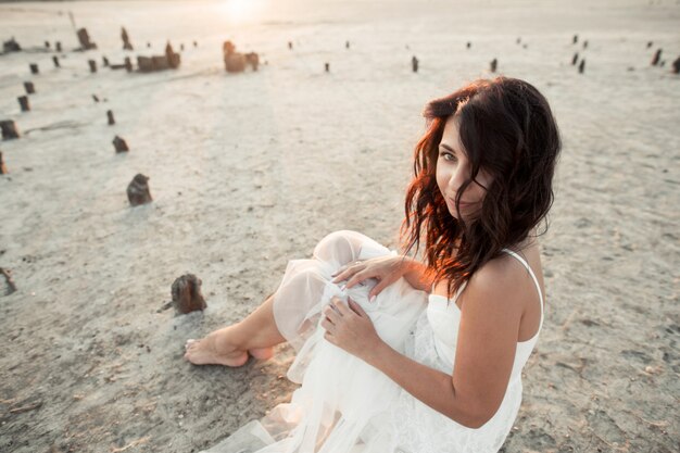 Young brunette caucasian girl is sitting on the sand, dressed in white dress and looking straight