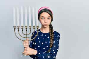 Free photo young brunette girl holding menorah hanukkah jewish candle puffing cheeks with funny face. mouth inflated with air, catching air.