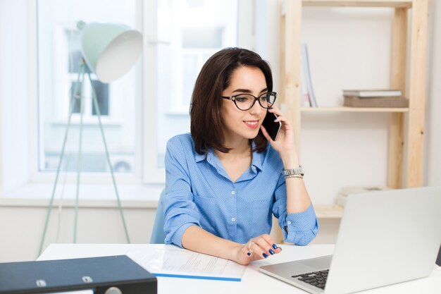 A young brunette girl is working at the table in office. She wears blue shirt and black glasses. She is speaking on phone.