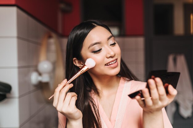 Young brunette woman applies makeup with brush and looks into small mirror