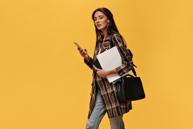 Young brunette woman in oversized jacket and jeans holds phone and white notebooks Attractive girl with black bag poses on orange background