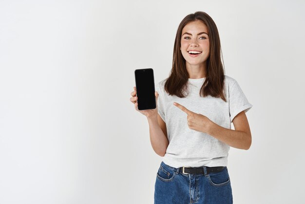 Young brunette woman showing blank smartphone screen and pointing finger at mobile phone smiling satisfied at camera standing over white background