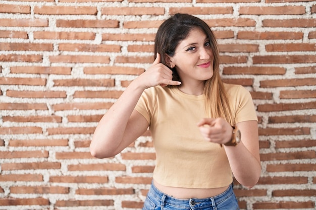 Young brunette woman standing over bricks wall smiling doing talking on the telephone gesture and pointing to you call me