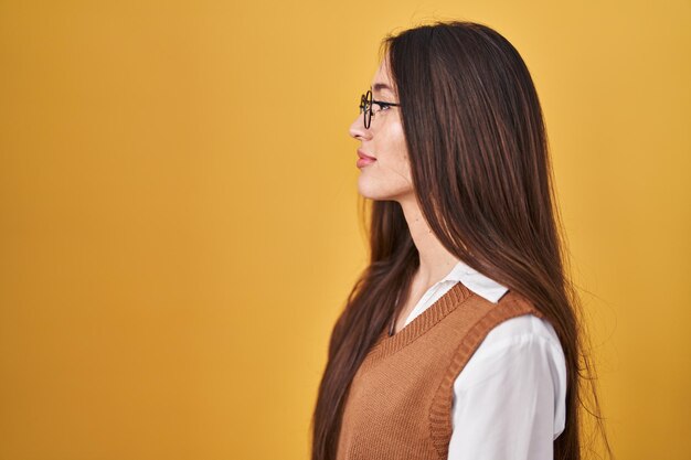 Young brunette woman standing over yellow background wearing glasses looking to side relax profile pose with natural face with confident smile