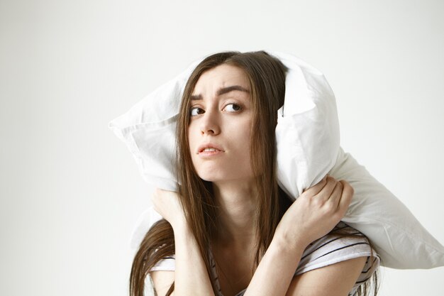 Free photo young brunette woman with messy hairstyle covering ears using white pillow looking sideways with frustration