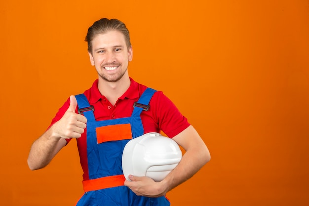 Young builder man in construction uniform holding safety helmet in hand and showing thumbs up with big smile on face standing over isolated orange wall with copy space