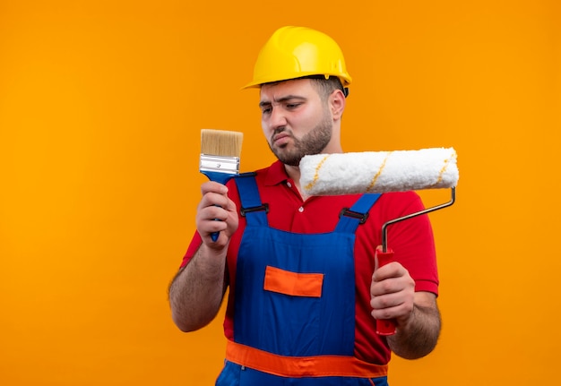 Free photo young builder man in construction uniform and safety helmet holding brush and paint roller looking at brush with skeptic expression