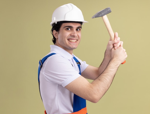 Young builder man in construction uniform and safety helmet holding hammer looking at front smiling slyly standing over green wall