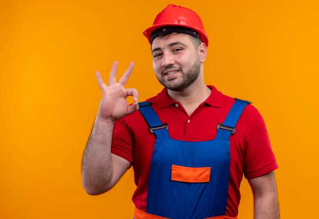 Young builder man in construction uniform and safety helmet looking at camera smiling cheerfully doing ok sign 