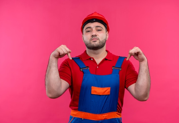 Free Photo young builder man in construction uniform and safety helmet looking confident pointing with index fingers down self-satisfied 