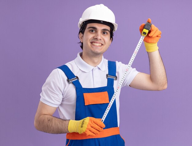 Young builder man in construction uniform and safety helmet in rubber gloves holding measure tape looking at front with smile on face standing over purple wall