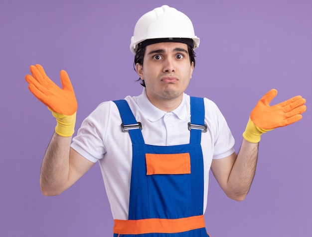 Young builder man in construction uniform and safety helmet in rubber gloves looking at front being confused and uncertain with arms raised standing over purple wall