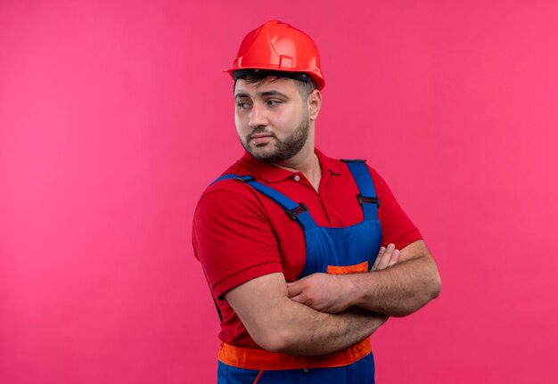 Free Photo young builder man in construction uniform and safety helmet  with hands on chest crossed looking aside with serious face