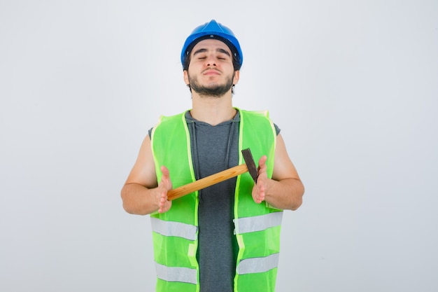 Free photo young builder man holding hammer while closing eyes in workwear uniform and looking confident , front view.