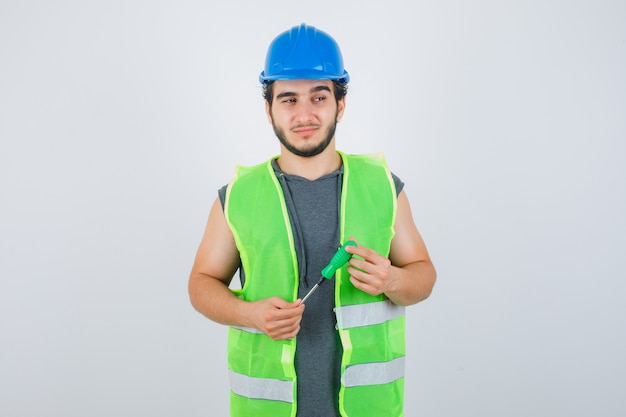 Free photo young builder man in uniform holding screwdriver while looking away and looking thoughtful , front view.