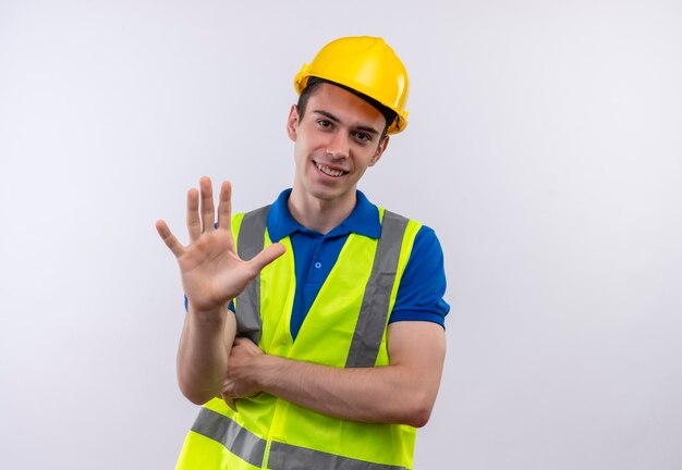 Young builder man wearing construction uniform and safety helmet happily shows five with hand