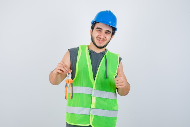 Young builder man in workwear uniform holding pliers while showing thumb up and looking pleased , front view.