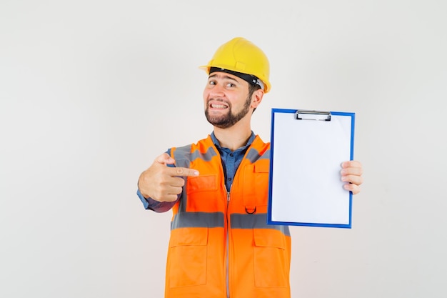 Free Photo young builder in shirt, vest, helmet pointing at clipboard and looking cheerful , front view.