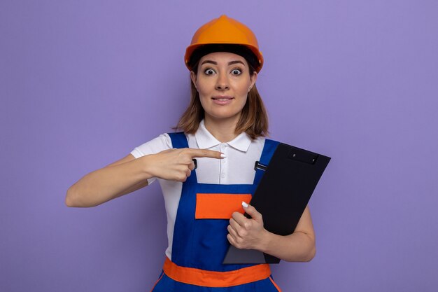 Young builder woman in construction uniform and safety helmet holding clipboard pointing with index finger at it  surprised and happy standing over purple wall