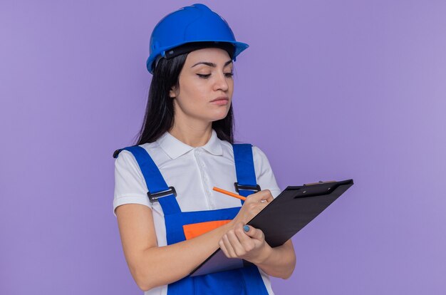 Young builder woman in construction uniform and safety helmet holding clipboard writing something looking confident standing over purple wall