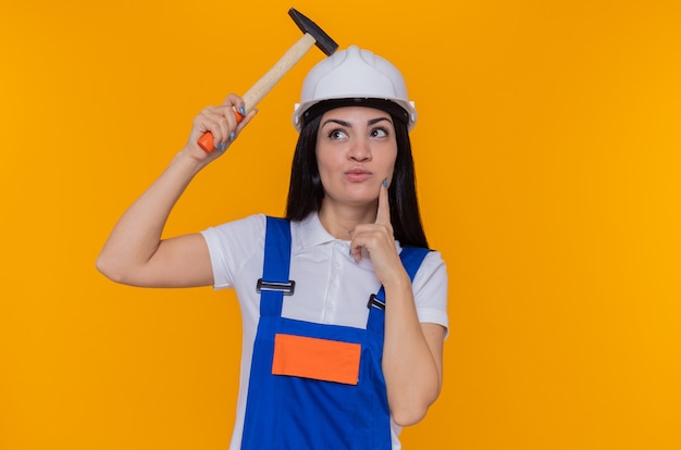 Young builder woman in construction uniform and safety helmet holding hammer looking up with pensive expression on face thinking standing over orange wall