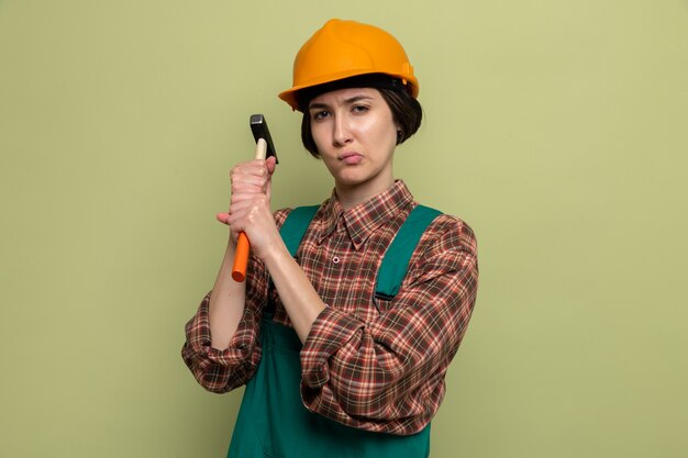 Young builder woman in construction uniform and safety helmet holding hammer with serious face standing on green