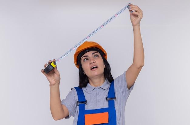 Free photo young builder woman in construction uniform and safety helmet holding measure tape looking at it with serious face standing over white wall