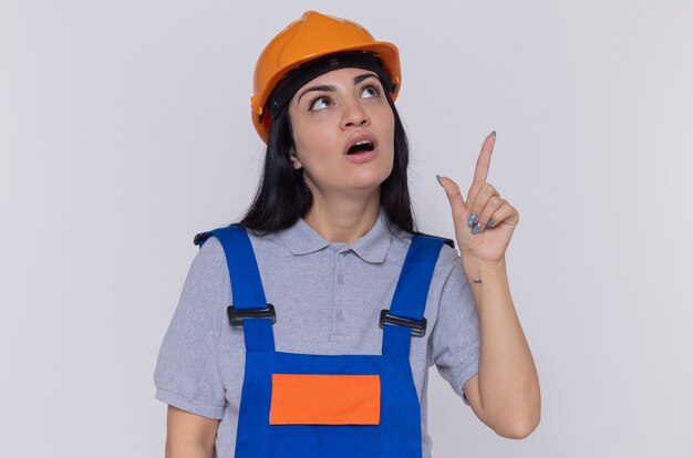 Young builder woman in construction uniform and safety helmet looking up surprised pointing with index finger at something standing over white wall