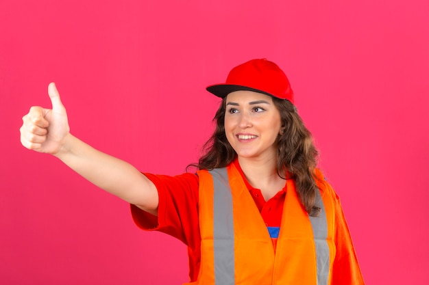 Young builder woman in construction uniform and safety helmet showing thumbs up to someone smiling friendly over isolated pink wall