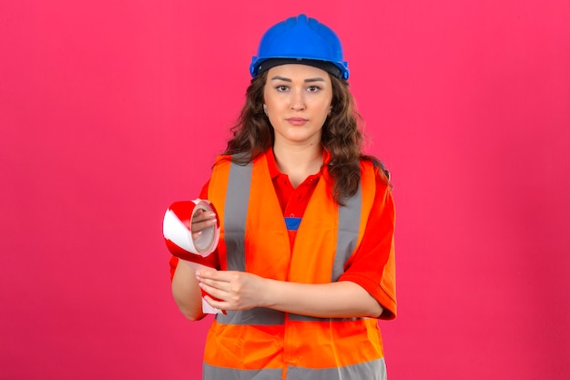 Young builder woman in construction uniform and safety helmet standing with adhesive tape with serious face over isolated pink wall_