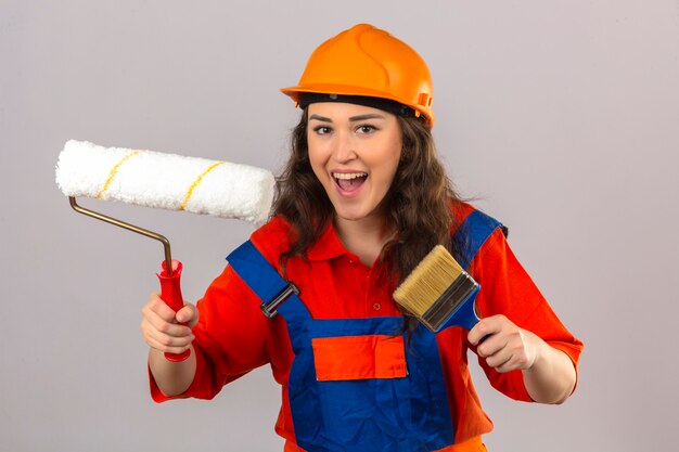 Young builder woman in construction uniform and safety helmet standing with paint roller and brush smiling cheerfully over isolated white wall