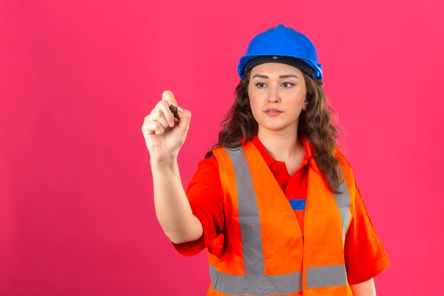 Young builder woman in construction uniform and safety helmet standing writing in air with pen over isolated pink wall