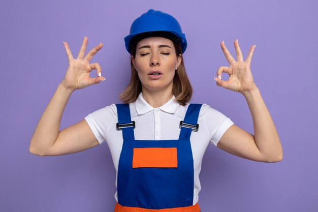 Young builder woman in construction uniform and safety helmet trying to relax making meditation gesture with fingers with eyes closed standing over purple wall