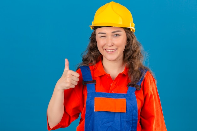 Young builder woman in construction uniform and safety helmet winking showing thumbs up with happy face over isolated blue wall