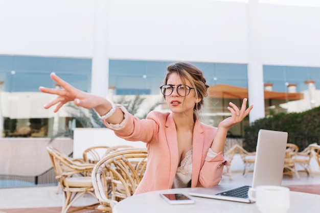 Free photo young business woman sitting in outdoor cafe with laptop on table, serious lady pointing with hand direction, saw something in afait. wearing stylish pink jacket, glasses, white watches.