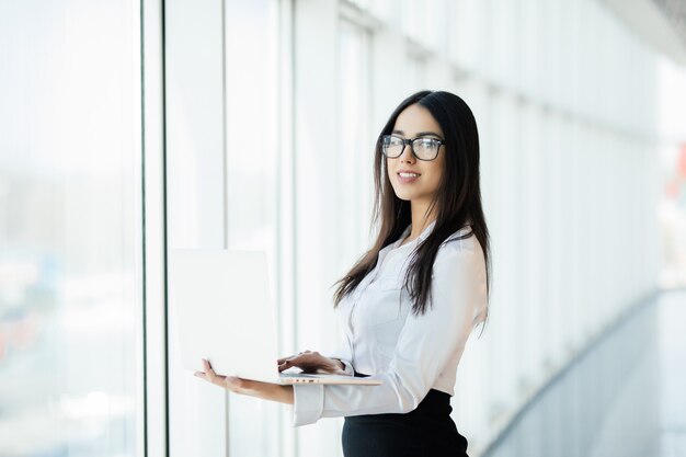 Young business woman working in her luxurious office holding a laptop standing against panoramic window with a view on business district