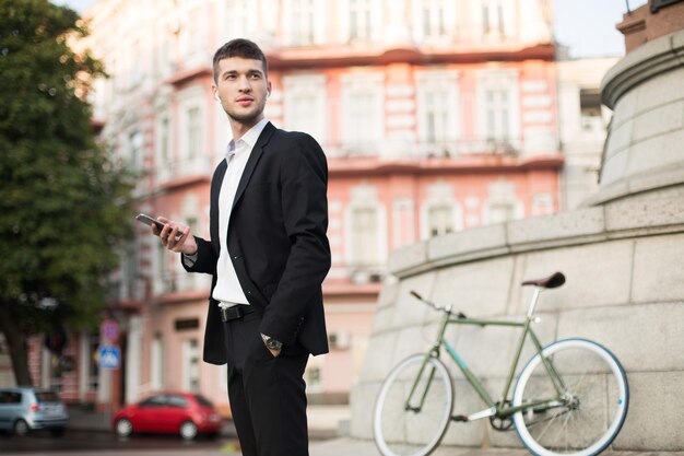 Young businessman in classic black suit and white shirt with wireless earphones holding cellphone in hand while thoughtfully looking aside with retro bicycle on background outdoor