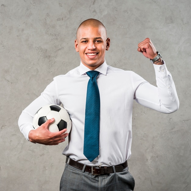 Free photo young businessman holding soccer ball in hand clenching her fist standing against grey wall