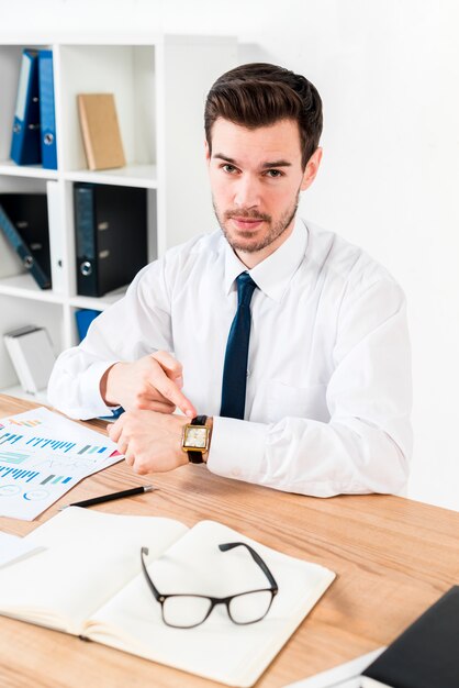 Young businessman looking to camera pointing his finger toward watch at workplace