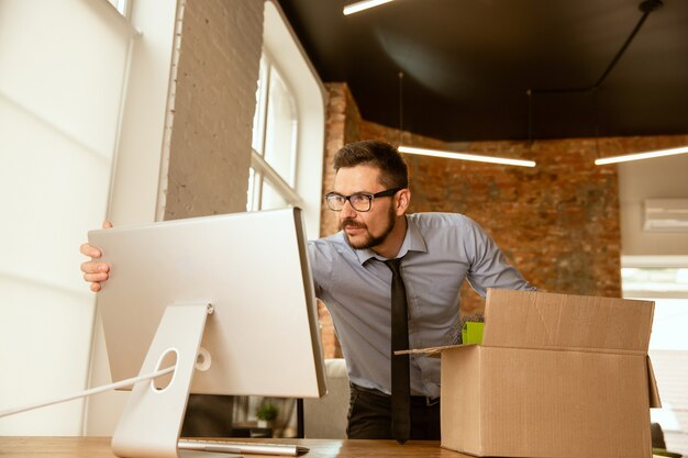 A young businessman moving in the office, getting new work place. Young caucasian male office worker equips new cabinet after promotion. Looks happy. Business, lifestyle, new life concept.