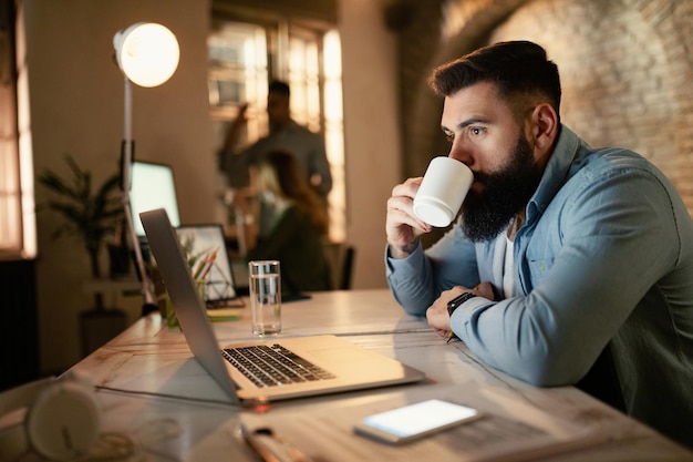 Young businessman surfing the net on a computer while drinking coffee in the evening at the office