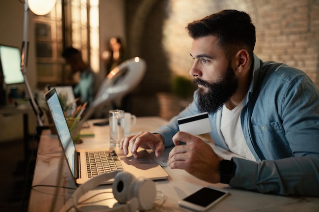 Young businessman using laptop and credit card while buying on the internet at night in the office