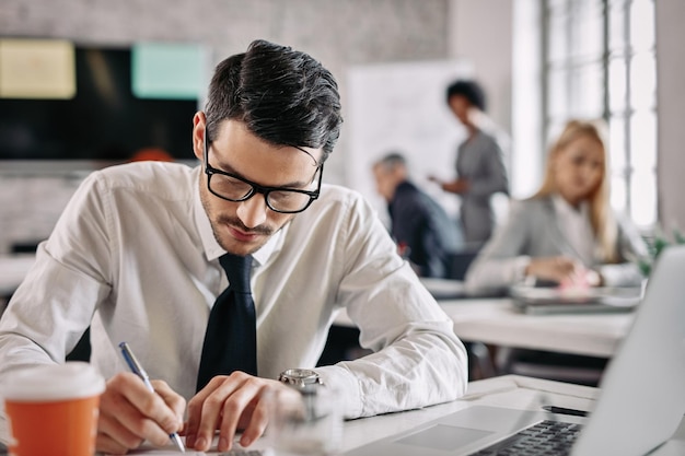 Young businessman writing at his desk while doing paperwork in the office There are people in the background