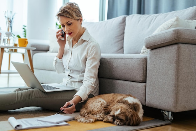 Young businesswoman communicating on mobile phone while analyzing report and using laptop at home