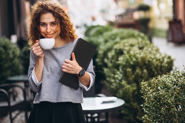 Free photo young businesswoman drinking coffee outside cafe holding laptop