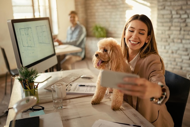 Young businesswoman having fun with her poodle while using phone and taking selfie in the office